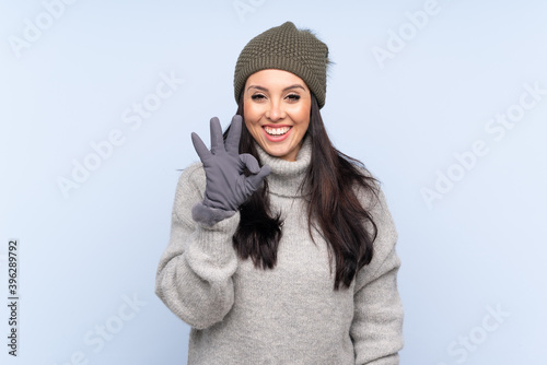 Young Colombian girl with winter hat over isolated blue background showing an ok sign with fingers