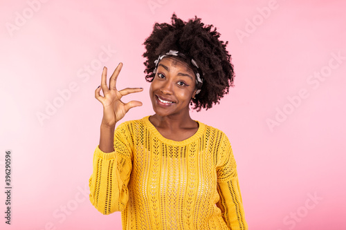Little bit. Pretty smiling black woman measures tiny invisble object, smiles gladfully, isolated on pink background, tells about salary income or decreased price