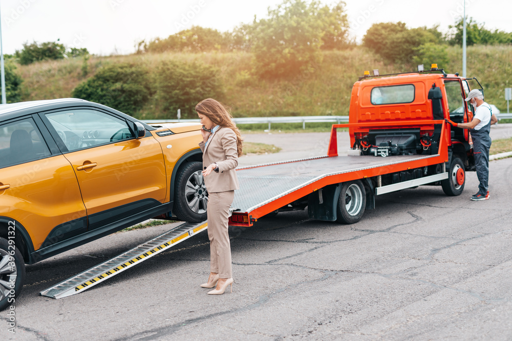 Elegant middle age business woman calling someone while towing service helping her on the road. Roadside assistance concept.