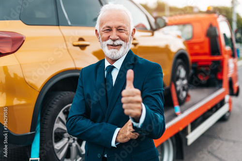 Elegant senior businesman is happy and satisfied with fast and reliable towing service for help on the road. He is standing in front of wrecker with arms crossed. Roadside assistance concept. © hedgehog94