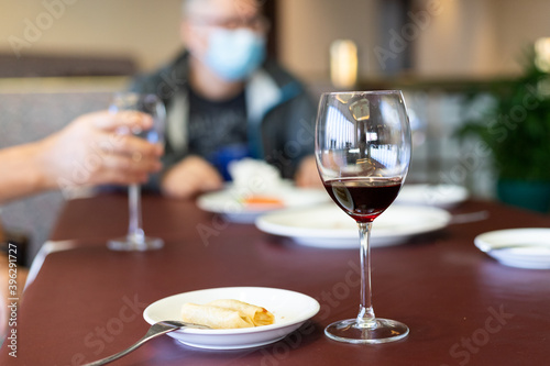 wine Glass and spring rolls on table, blurred person with face mask in background. Gathering in bar or restaurant limited to 4 people, leading to bad business. face mask mandatory in public settings
