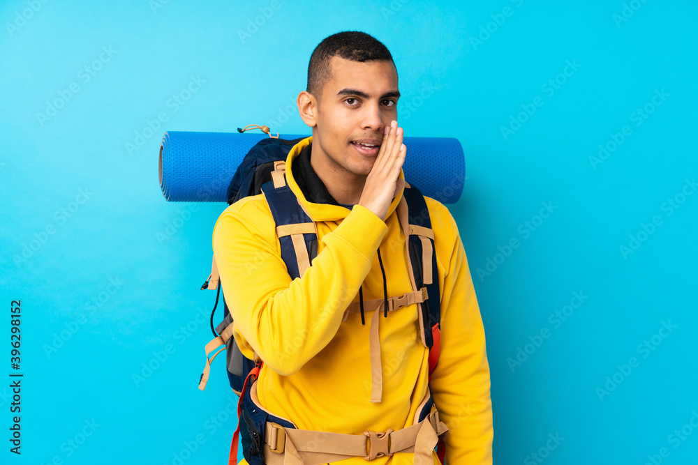 Young mountaineer man with a big backpack over isolated blue background whispering something Young mountaineer man with a big backpack over isolated blue background whispering something
