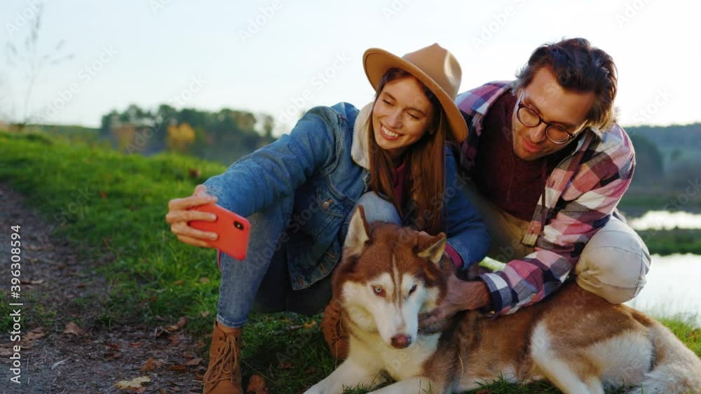 Lovely couple with a dog taking selfie photo on phone in autumn forest near lake. Landscape vacation relationship pets. Outdoors nature. Slow motion Lovely couple with a dog taking selfie photo on phone in autumn forest near lake. Landscape vacation relationship pets. Outdoors nature. Slow motion