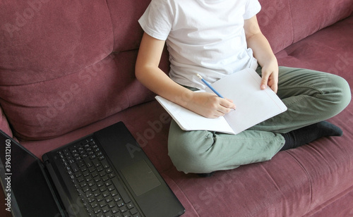 The child is at home, on the couch, studying on the computer online and making notes in a notebook.