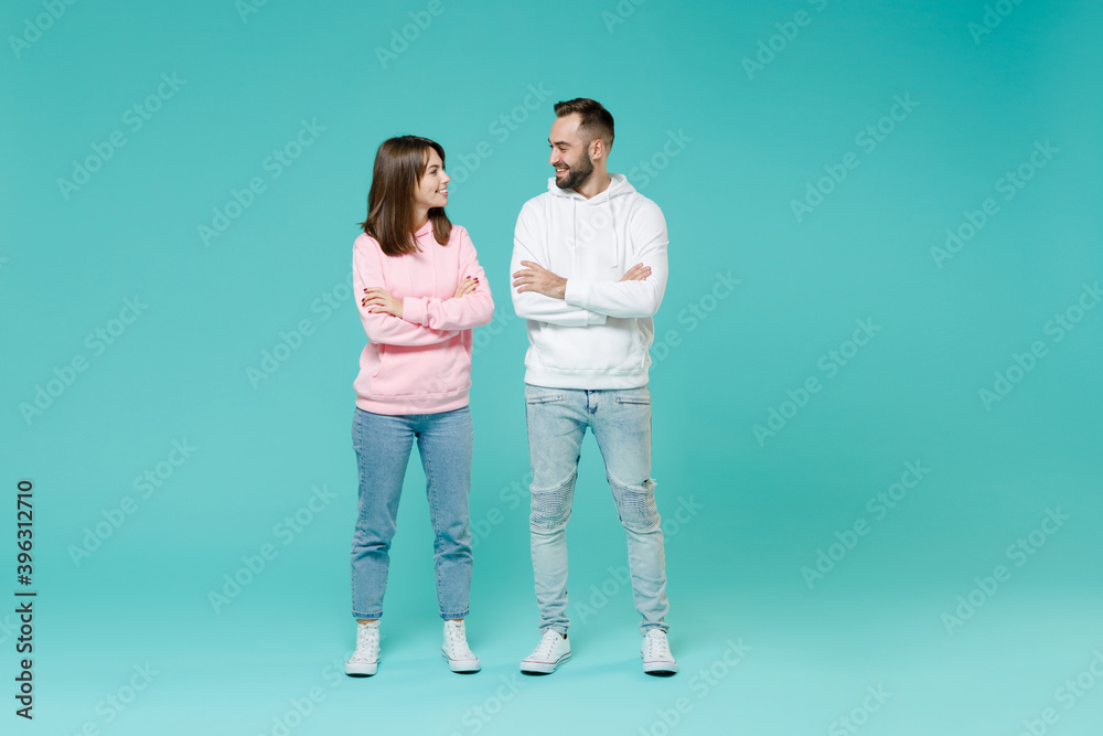 Full length of smiling young couple two friends man woman 20s wearing white pink casual hoodie holding hands crossed looking at each other isolated on blue turquoise wall background studio portrait. Full length of smiling young couple two friends man woman 20s wearing white pink casual hoodie holding hands crossed looking at each other isolated on blue turquoise wall background studio portrait.