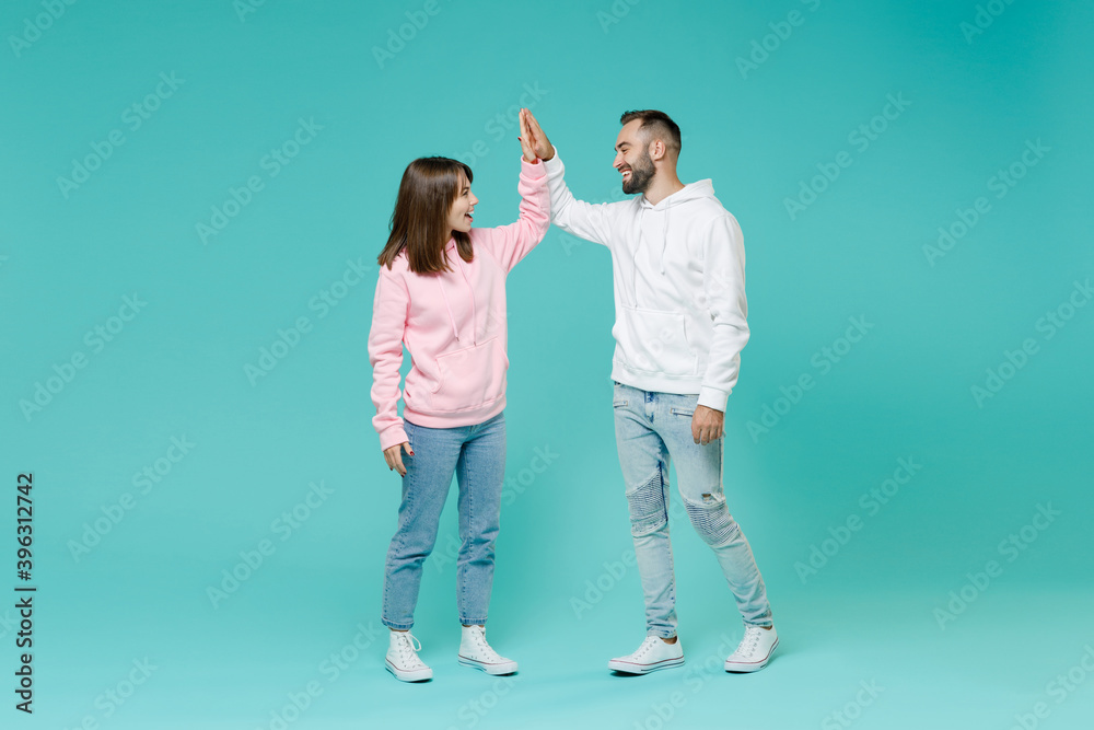 Full length of cheerful young couple two friends man woman in white pink casual hoodie hold hands folded giving high five looking at each other isolated on blue turquoise background studio portrait.