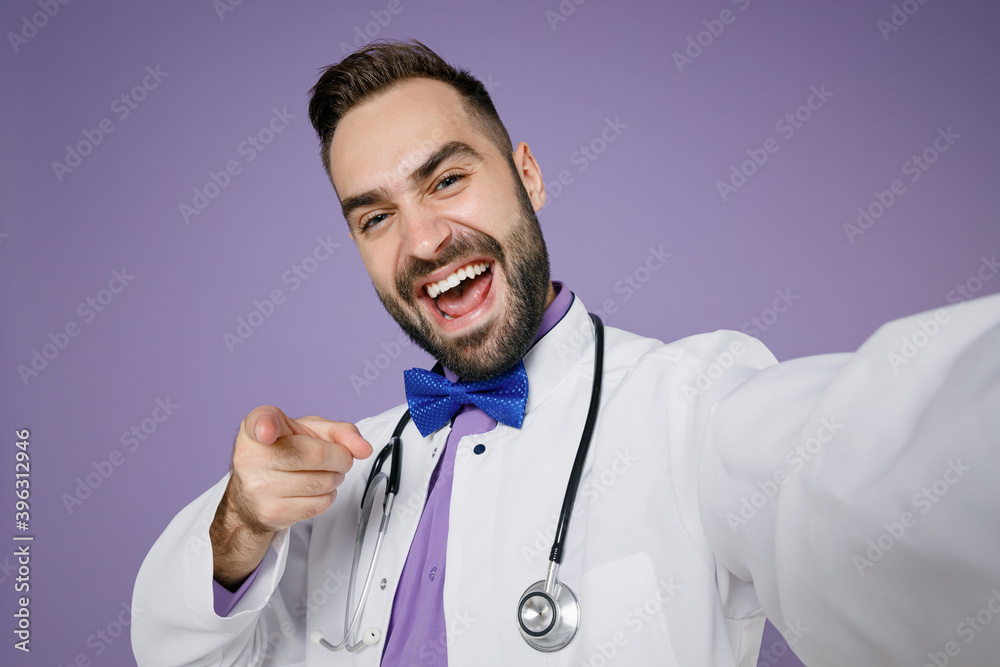 Close up of funny young bearded doctor man in white medical gown doing  selfie shot on mobile phone point finger on camera isolated on violet  background. Healthcare personnel health medicine concept. Stock