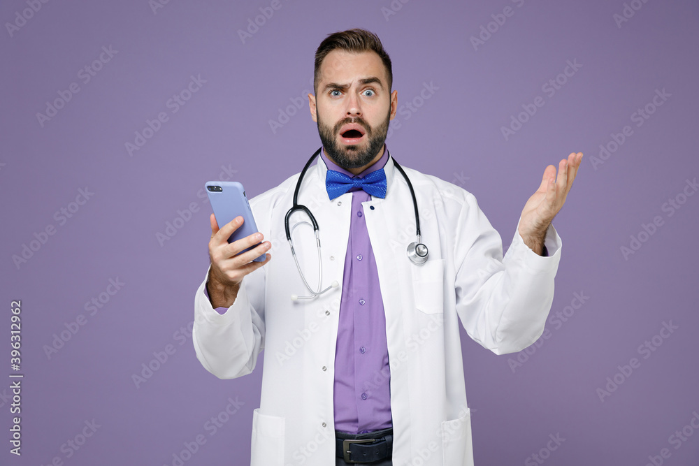 Shocked worried young bearded doctor man in medical gown stethoscope using mobile phone typing sms message spreading hands isolated on violet background. Healthcare personnel health medicine concept. Shocked worried young bearded doctor man in medical gown stethoscope using mobile phone typing sms message spreading hands isolated on violet background. Healthcare personnel health medicine concept.