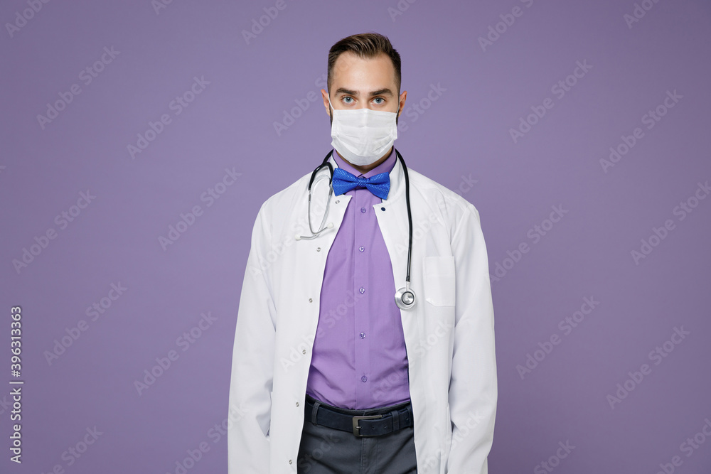 Young bearded doctor man wearing white medical gown stethoscope sterile face mask to safe from coronavirus virus covid-19 isolated on violet background. Healthcare personnel health medicine concept. Young bearded doctor man wearing white medical gown stethoscope sterile face mask to safe from coronavirus virus covid-19 isolated on violet background. Healthcare personnel health medicine concept.