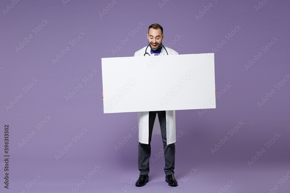 Full length of excited young bearded doctor man in white medical gown hold big white empty blank billboard isolated on violet background studio portrait. Healthcare personnel health medicine concept. Full length of excited young bearded doctor man in white medical gown hold big white empty blank billboard isolated on violet background studio portrait. Healthcare personnel health medicine concept.