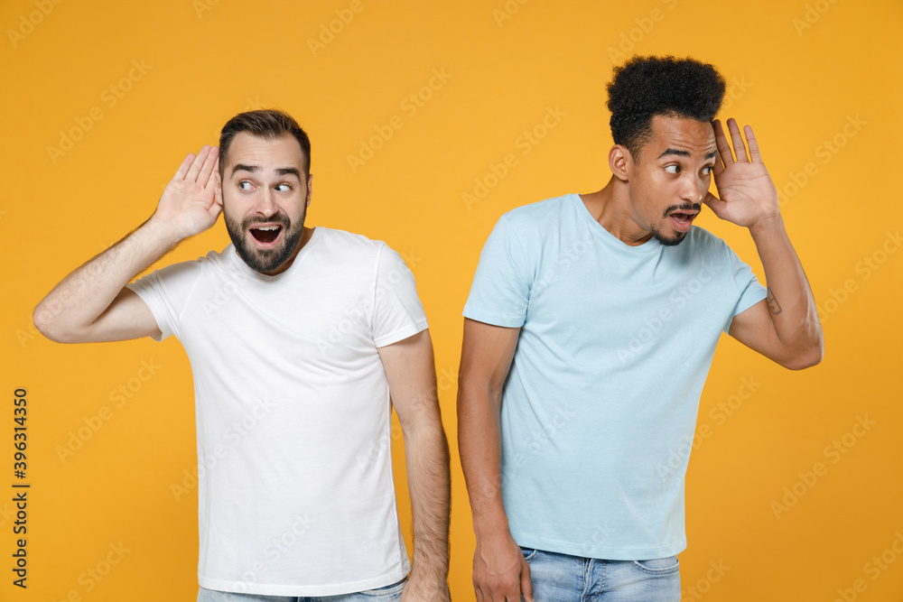 Curious young two friends european african american men 20s in white blue casual t-shirts try to hear you overhear listen intently looking aside isolated on yellow colour background studio portrait. Curious young two friends european african american men 20s in white blue casual t-shirts try to hear you overhear listen intently looking aside isolated on yellow colour background studio portrait.