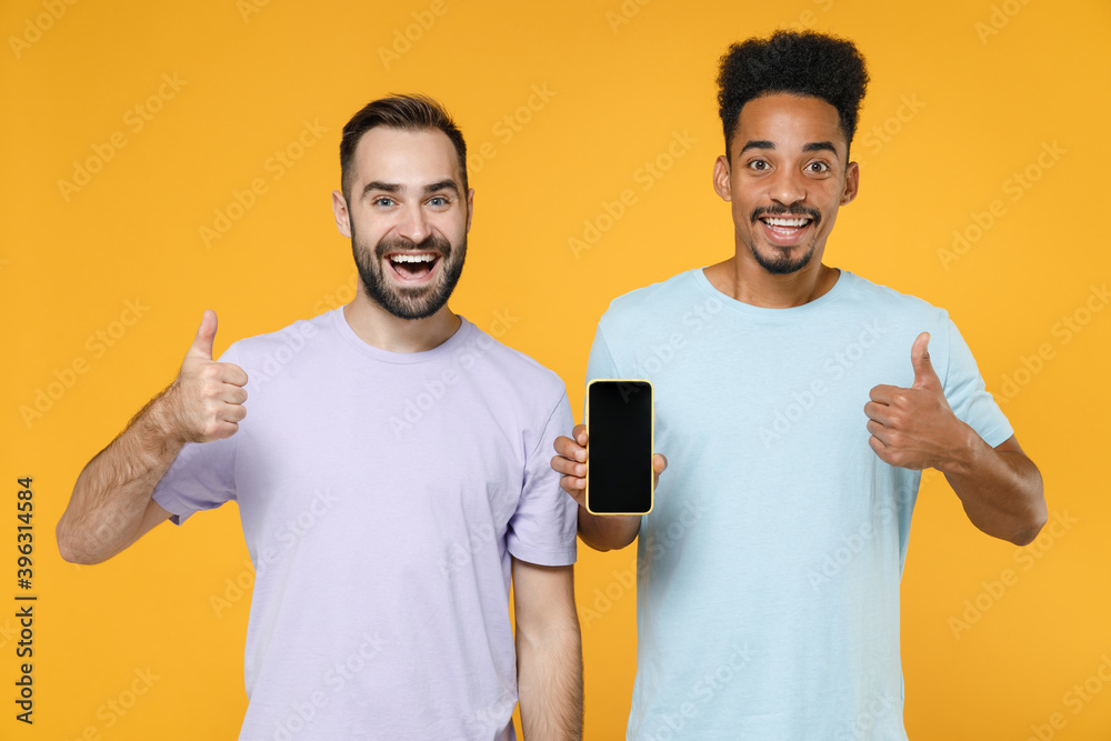 Excited young two friends european african american men 20s in violet blue t-shirts using mobile cell phone with blank empty screen showing thumb up isolated on yellow background studio portrait.