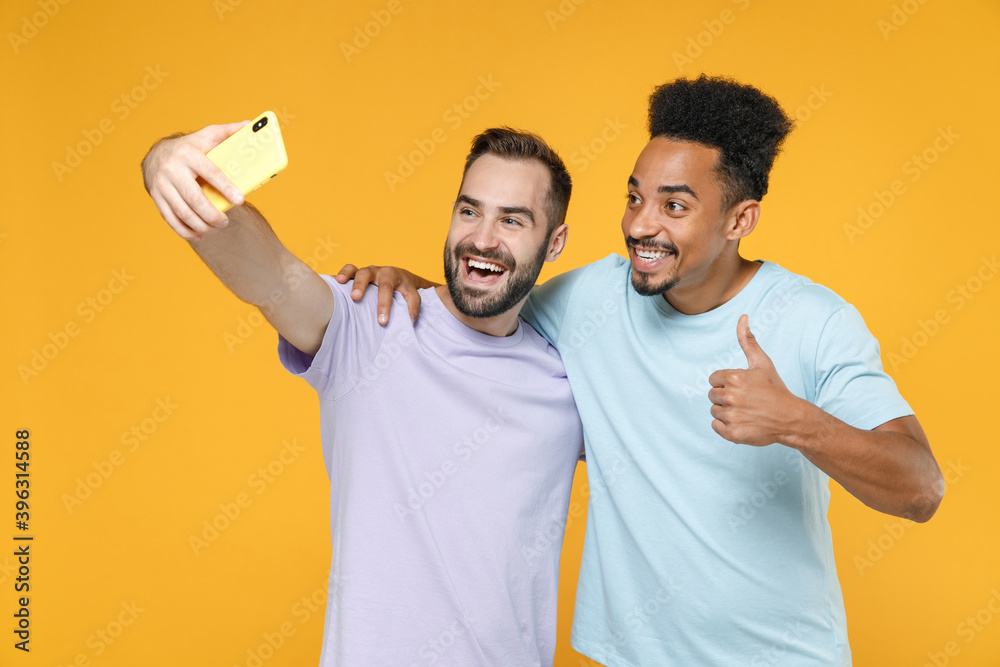 Funny young two friends european african american men 20s wearing violet blue casual t-shirts doing selfie shot on mobile phone showing thumb up isolated on yellow colour background studio portrait.