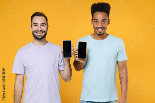 Smiling young two friends european african american men 20s in violet blue casual t-shirts using mobile cell phone with blank empty screen isolated on bright yellow colour background studio portrait.