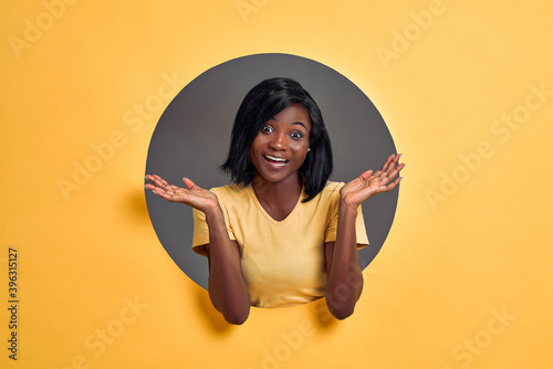 Unsure afro girl shrugging shoulders with spreaded hands and opened empty palms. Woman in a round hole circle in orange background. Copy space. © HBS