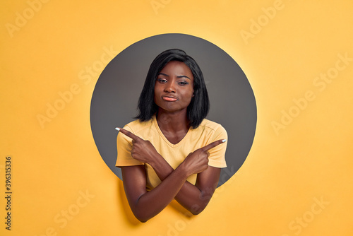 Young woman standing wearing casual t-shirt in a round circle hole in orange background pointing to both sides with fingers, different direction disagree. Copy space. © HBS