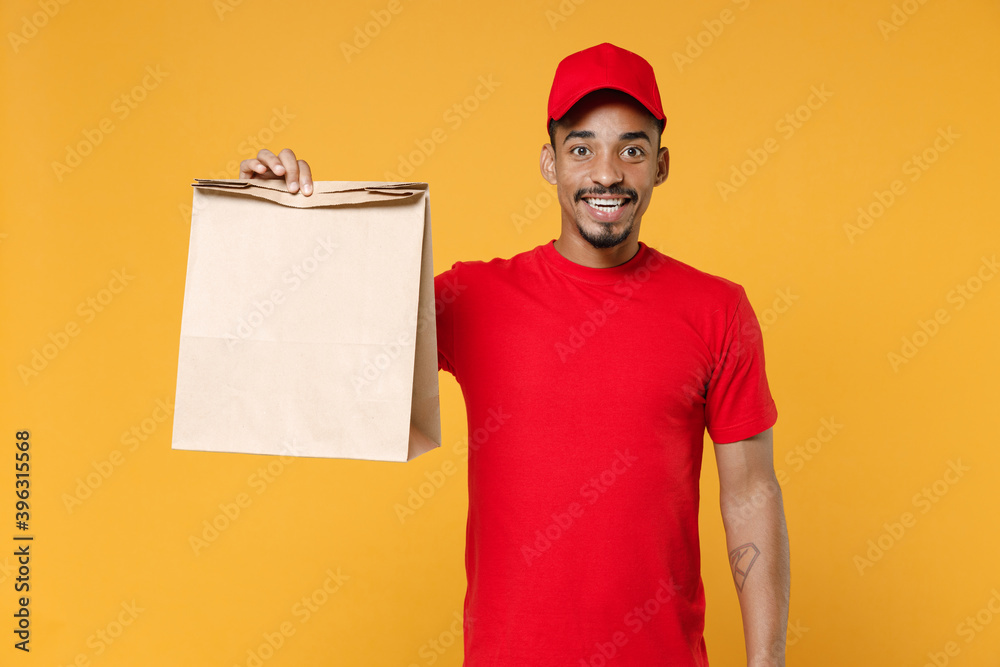 Delivery employee african man 20s in red cap blank print t-shirt uniform work courier dealer service hold brown clear empty craft paper bag for takeaway mock up isolated on yellow background studio