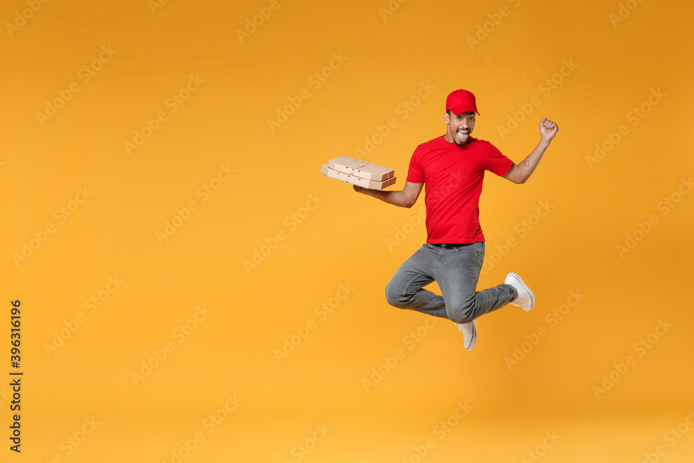 Full length delivery employee african man in red cap blank print t-shirt uniform work courier service concept hold give food order pizza cardboard boxes jump run isolated on yellow background studio.