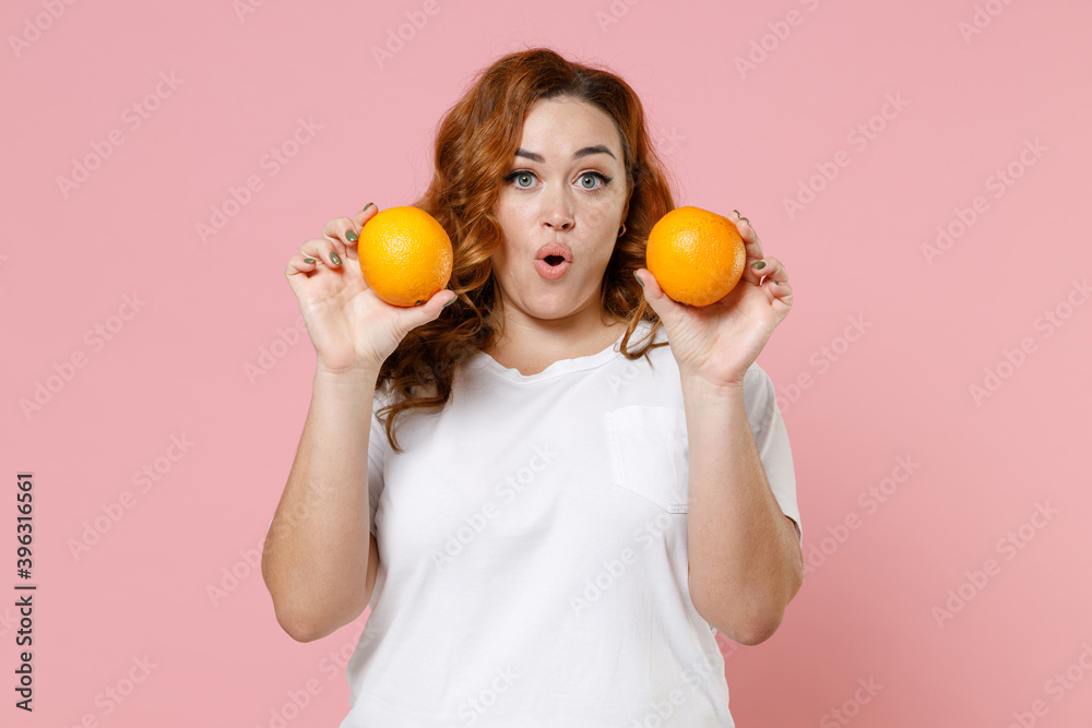 Shocked amazed young redhead plus size body positive woman 20s wearing white casual t-shirt hold ripe fresh orange fruits looking camera isolated on pastel pink color wall background studio portrait. Shocked amazed young redhead plus size body positive woman 20s wearing white casual t-shirt hold ripe fresh orange fruits looking camera isolated on pastel pink color wall background studio portrait.