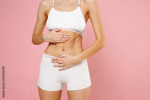 Cropped image of beautiful seductive young woman wearing white brassiere underwear with sports fit body standing posing hold hands on stomach belly isolated on pastel pink background, studio portrait. photo