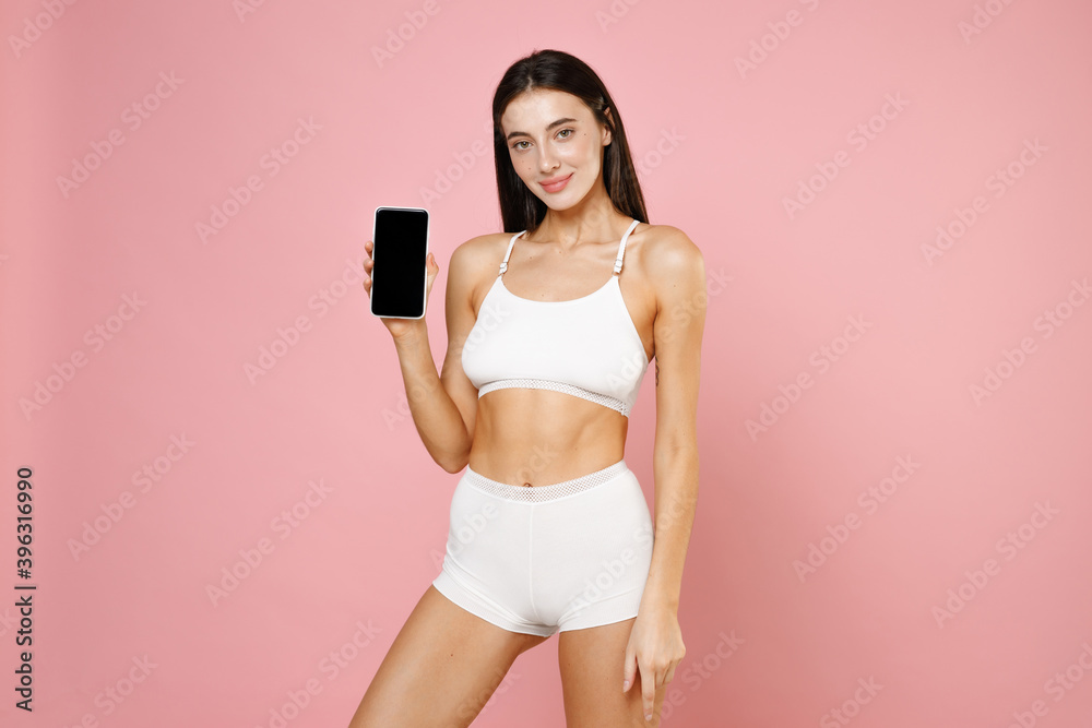 Smiling beautiful charming young brunette woman 20s in white underwear posing hold mobile cell phone with blank empty screen looking camera isolated on pastel pink colour background studio portrait. Smiling beautiful charming young brunette woman 20s in white underwear posing hold mobile cell phone with blank empty screen looking camera isolated on pastel pink colour background studio portrait.