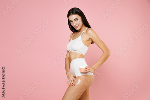 Side view of smiling attractive sexy young brunette woman 20s in white underwear posing applying moisturizer body cream from container isolated on pastel pink colour wall background, studio portrait. © ViDi Studio