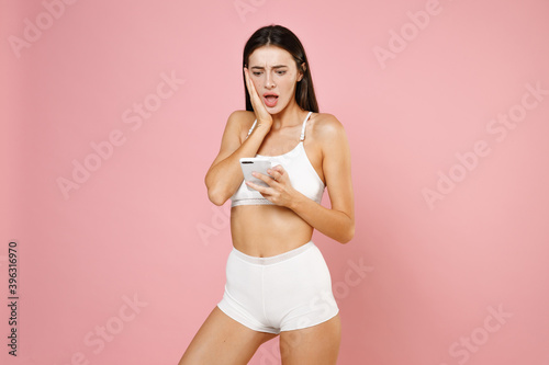 Shocked amazed worried young brunette woman 20s wearing white underwear posing using mobile cell phone typing sms message put hand on cheek isolated on pastel pink colour background studio portrait.