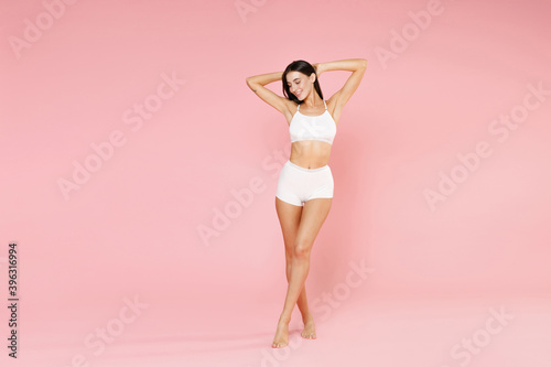 Full length of smiling young brunette woman in white underwear standing with hands up showing result on underarms removing hair procedure isolated on pastel pink colour background, studio portrait.