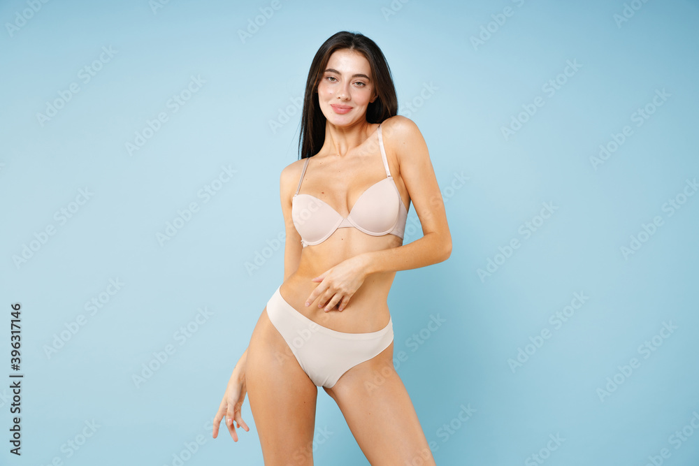 Smiling pretty young brunette woman 20s in beige underwear showing sports  body standing posing hold hand on stomach belly looking camera isolated on  pastel blue colour background studio portrait. Stock Photo