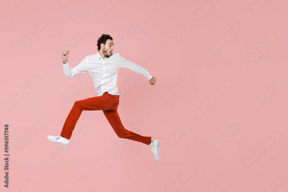 Full length side view of shocked worried young bearded man 20s wearing basic casual white shirt jumping like running looking aside isolated on pastel pink color wall background studio portrait.