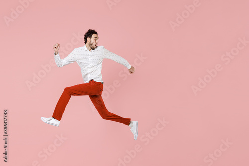 Full length side view of shocked worried young bearded man 20s wearing basic casual white shirt jumping like running looking aside isolated on pastel pink color wall background studio portrait. © ViDi Studio