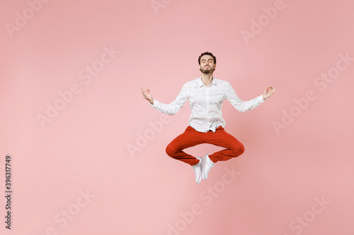 Full length of young bearded man 20s in basic casual white shirt jumping hold hands in yoga gesture, relaxing meditating, trying to calm down isolated on pastel pink color background studio portrait.
