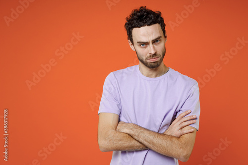 Displeased dissatisfied concerned young bearded man 20s wearing casual violet t-shirt standing holding hands crossed looking camera isolated on bright orange color wall background studio portrait.