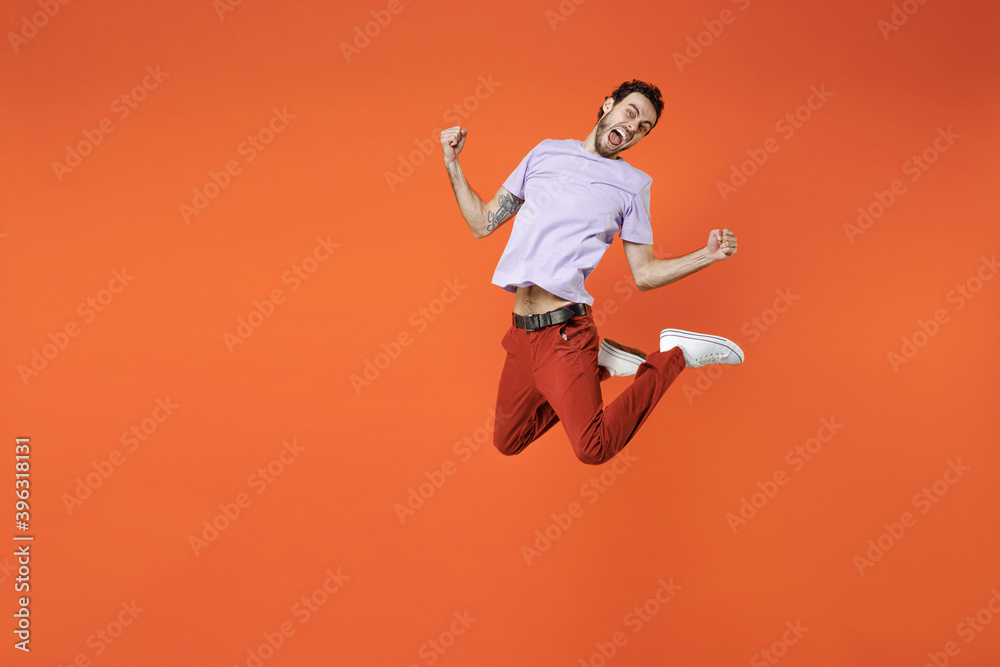 Full length of overjoyed screaming young bearded man 20s wearing casual violet t-shirt jumping doing winner gesture clenching fists looking camera isolated on bright orange background studio portrait.
