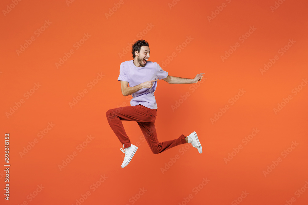 Full length side view of excited screaming young bearded man 20s wearing casual violet t-shirt jumping pointing index fingers aside isolated on bright orange color wall background studio portrait. Full length side view of excited screaming young bearded man 20s wearing casual violet t-shirt jumping pointing index fingers aside isolated on bright orange color wall background studio portrait.
