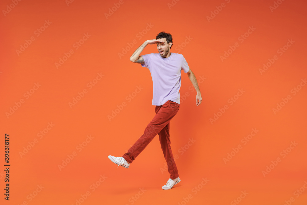 Full length of excited cheerful young bearded man 20s in casual violet t-shirt standing holding hand at forehead looking far away distance isolated on bright orange color background studio portrait.