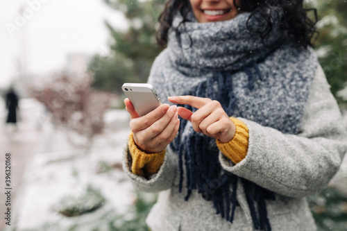 Young woman using her mobile phone at a snowy winter park. Closeup of female checking news and texting on her cellphone outdoor during cold winter season. Peoples' gadgets concept.