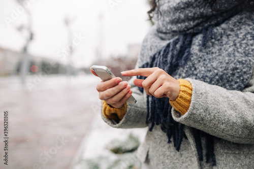 Young woman using her mobile phone at a snowy winter park. Closeup of female checking news and texting on her cellphone outdoor during cold winter season. Peoples' gadgets concept.
