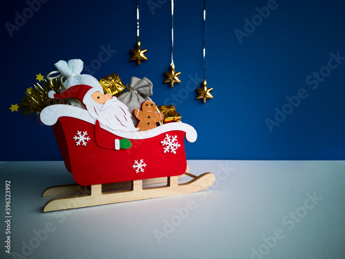 Christmas gift concept.Santa Claus in red sleigh on wooden runners with Ginger,gray,white sackfuls of gifts,gold box,on white blue blurred background,golden stars decor on silver tinsel.Copy space photo