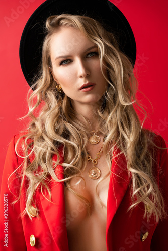 A beautiful blonde curly girl wearing a red pantsuit, unbuttoned on her braless big breasts and hat on a red background. Advertising, fashionable, commercial, closeup design