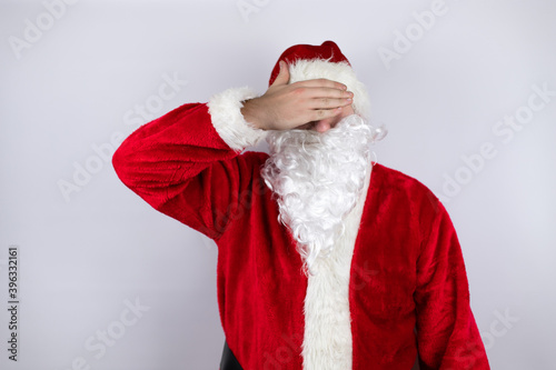 Man dressed as Santa Claus standing over isolated white background serious and covering her eyes with her hand