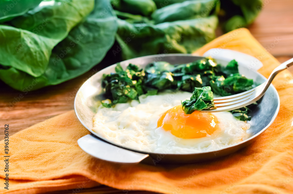 Green cooked spinach with fried egg on iron pan plate. Fried eggs healthy breakfast eating on wooden table background served by orange napkin, fork and spinach leaves, ingredients. Close up Green cooked spinach with fried egg on iron pan plate. Fried eggs healthy breakfast eating on wooden table background served by orange napkin, fork and spinach leaves, ingredients. Close up