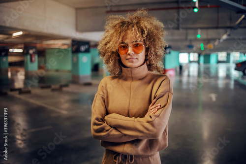 Obraz na plátne Young fashionable woman with curly hair standing in underground garage with arms crossed and looking at camera