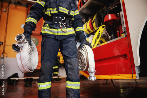 Fototapeta Firefighter in protective uniform with helmet on head checking on hoses before intervention while standing in fire station