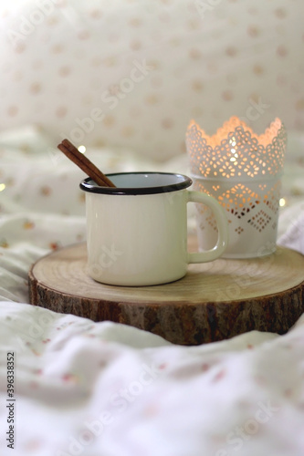 Enamel mug with warm drink and cinnamon stick and lit candle. Selective focus.