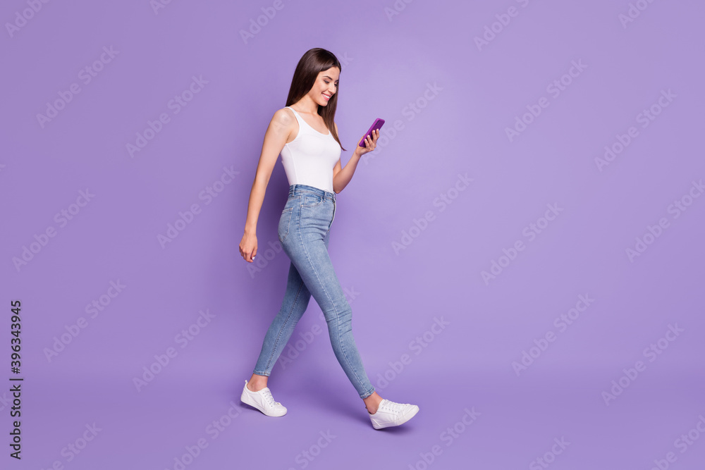 Full size profle side photo of happy brown haired girl text walk hold phone wear white tank-top isolated on purple color background Full size profle side photo of happy brown haired girl text walk hold phone wear white tank-top isolated on purple color background