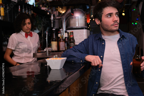Young caucasian man with a beer bottle in his hand watching TV, football match in a bar. Waitress on the background.
