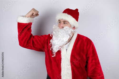 Man dressed as Santa Claus standing over isolated white background showing arms muscles smiling proud