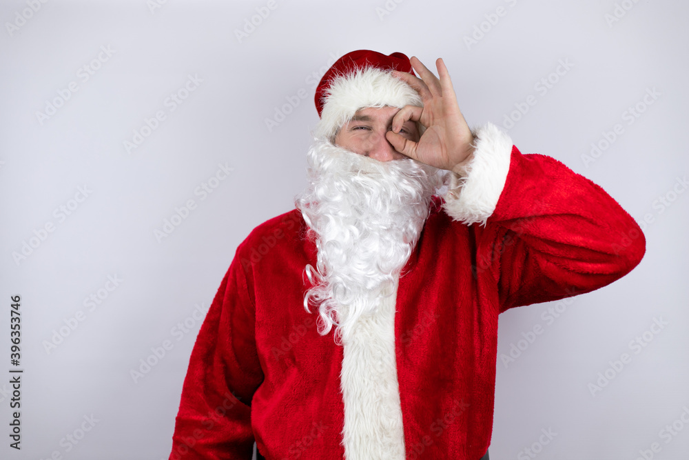 Man dressed as Santa Claus standing over isolated white background doing ok gesture shocked with smiling face, eye looking through fingers Man dressed as Santa Claus standing over isolated white background doing ok gesture shocked with smiling face, eye looking through fingers