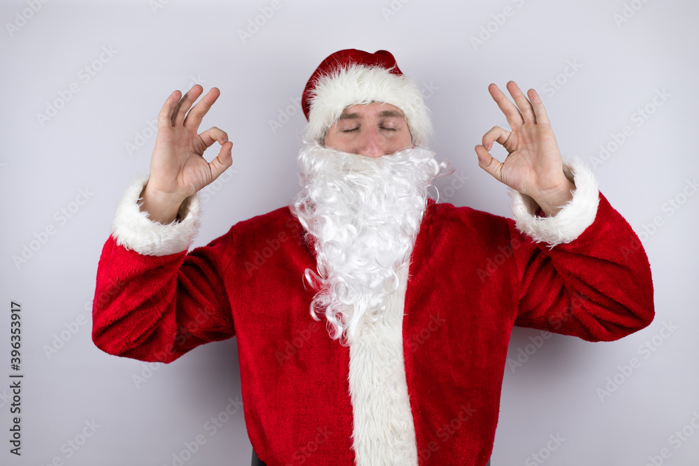 Man dressed as Santa Claus standing over isolated white background relax and smiling with eyes closed doing meditation gesture with fingers Man dressed as Santa Claus standing over isolated white background relax and smiling with eyes closed doing meditation gesture with fingers
